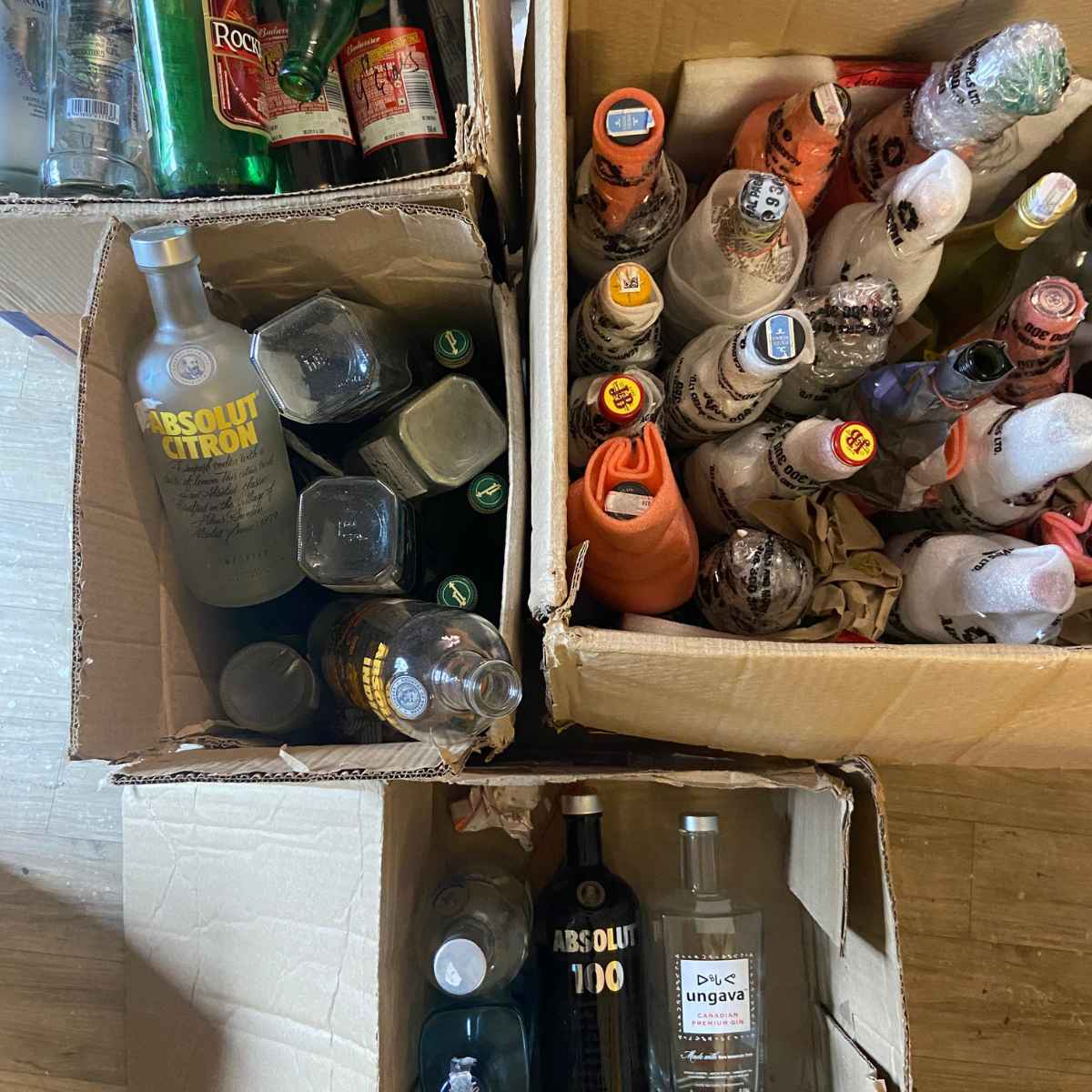 Three cardboard boxes filled with assorted liquor and spirit bottles, including visible Absolut Citron, Absolut 100, and other branded bottles, photographed from above on a wood floor.
